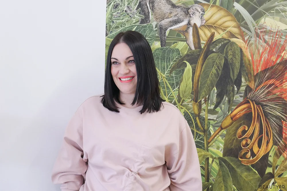 Dar haired woman in bright orange lipstick and blush pink sweater in front of a leafy jungle background