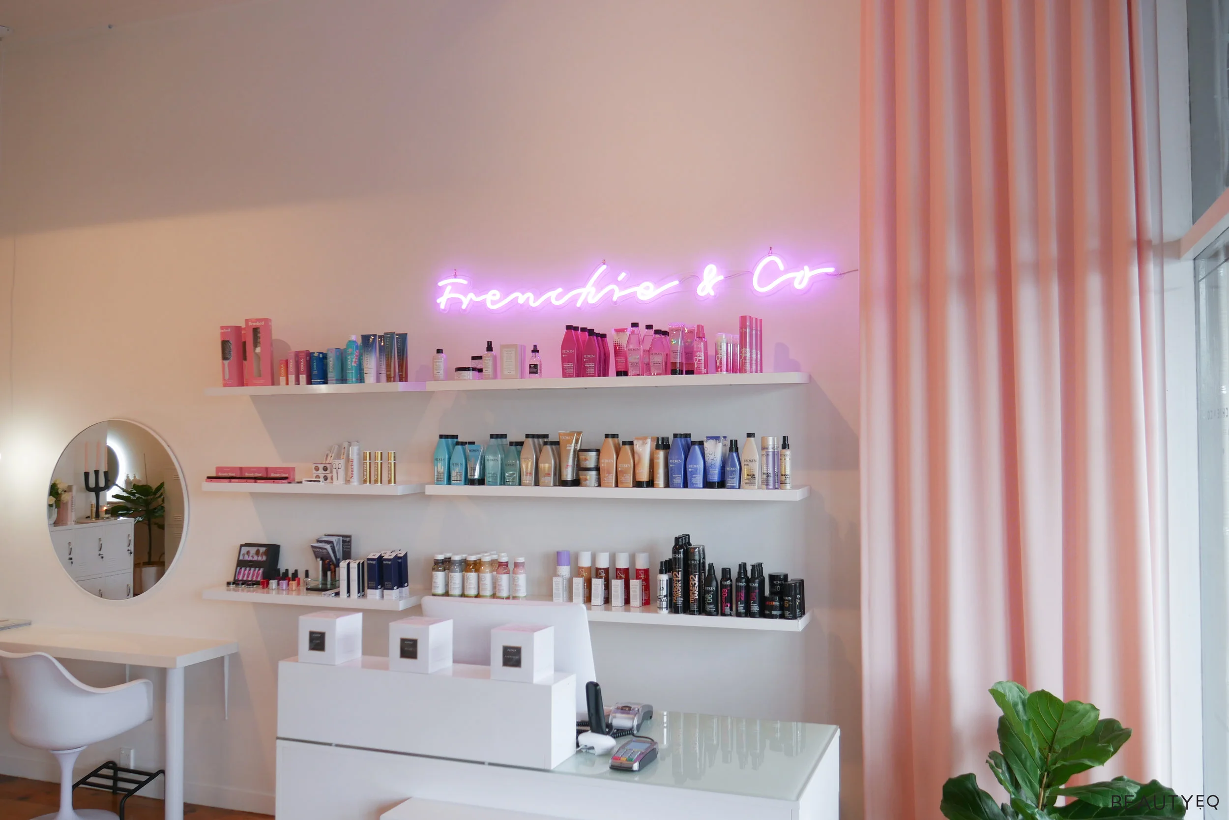 A beauty salon with various beauty products lines up on three different shelves and a neon light above them saying 'Frenchie & Co'