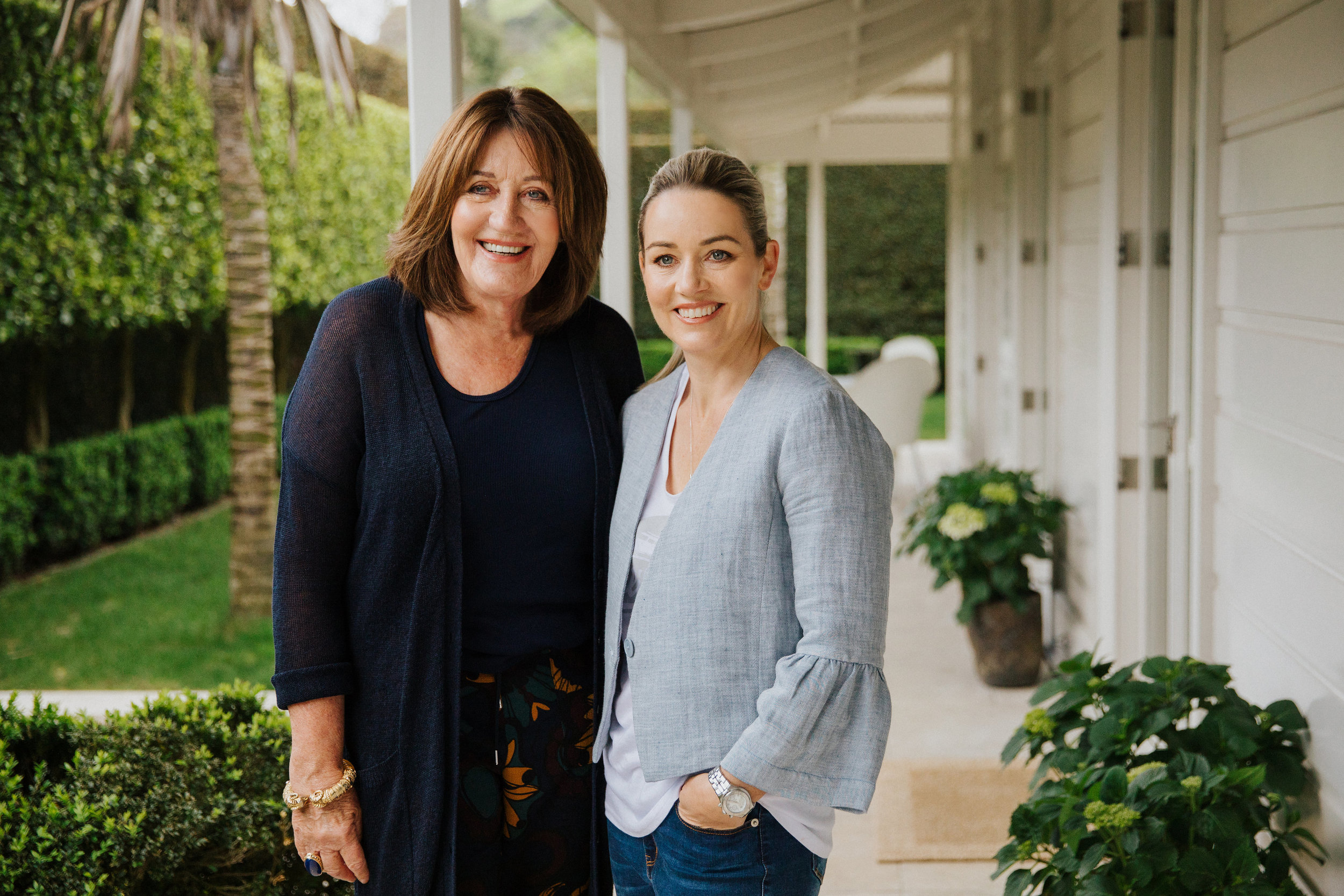 Two woman standing next to each other in the porch area if a house with hands in pockets