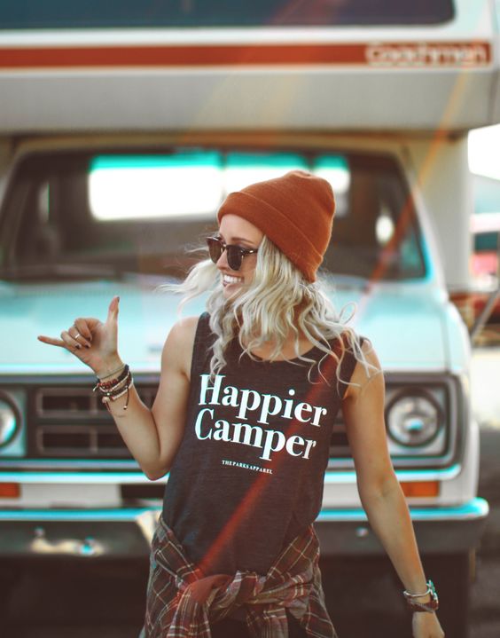 A blonde girl wearing an orange beanie and 'Happier Camper' tank top with a plaid shirt wrapped around her waist, pulling the 'shakka' sign infront of a blue car 