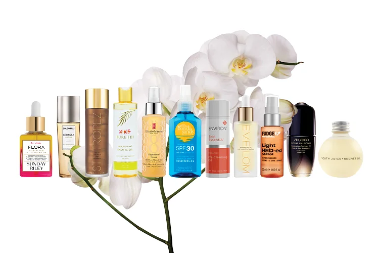 A range of different beauty oils lined up side by side against a white background with white flowers on skinny stems behind them 