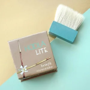 A Hoola light makeup product laid out on a blue and yellow background next to a brush 