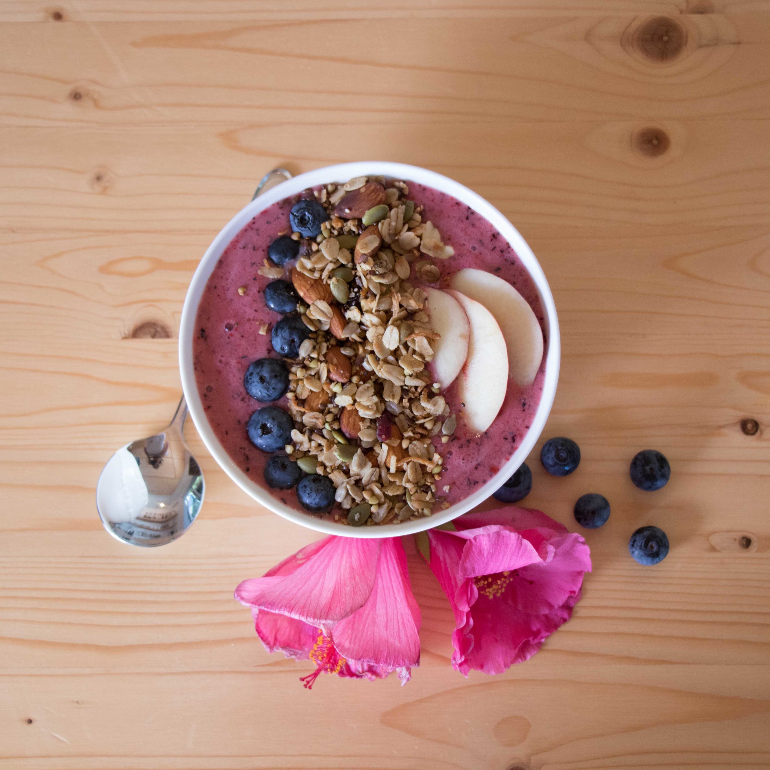 Smoothie bowl on wooden table next to pink flowers 