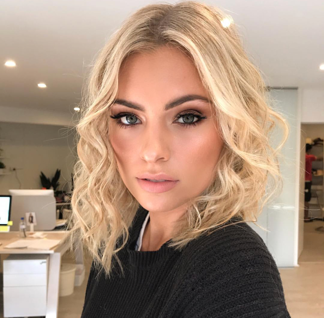 A blonde female model with curled, shoulder length hair and dark eye brows, wearing black knit jumper 
