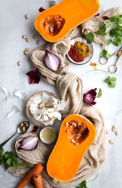 A range of vegetables such as onions, garlic, squash, some coriander and seeds laid out on a white background with against a long cloth 