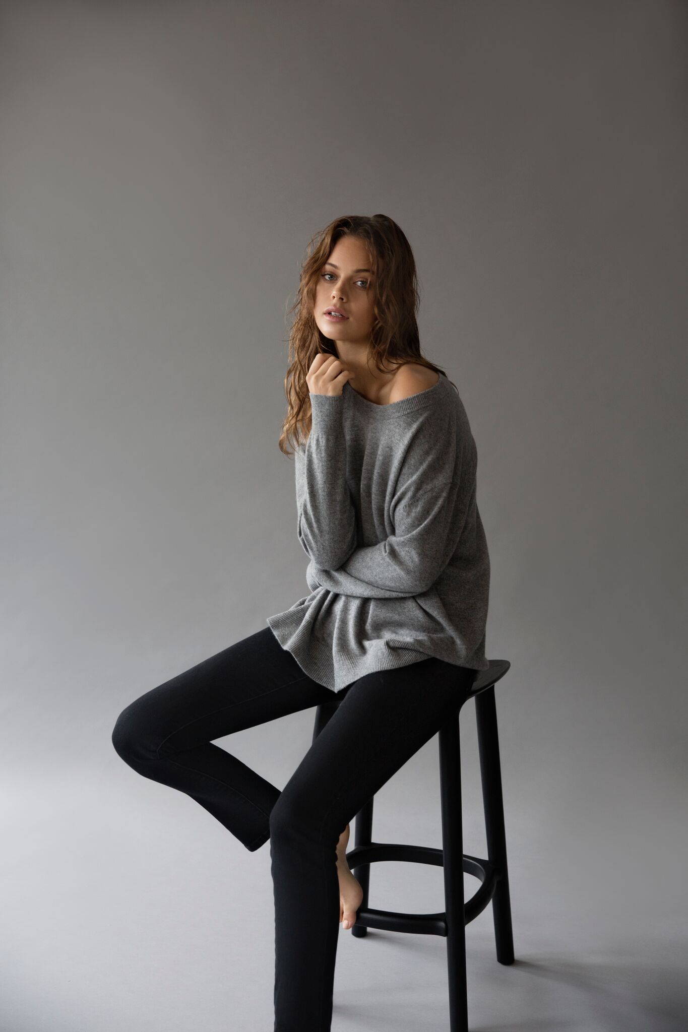 Brunette model in grey long sleeve sitting on black stool, against grey background