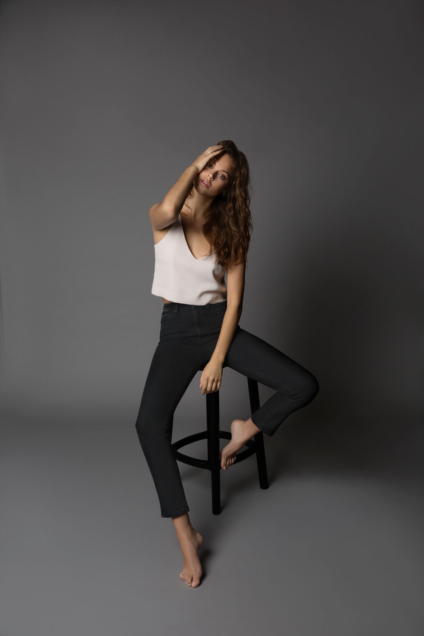 Brunette model in white top and black pants sitting on stool against white background 