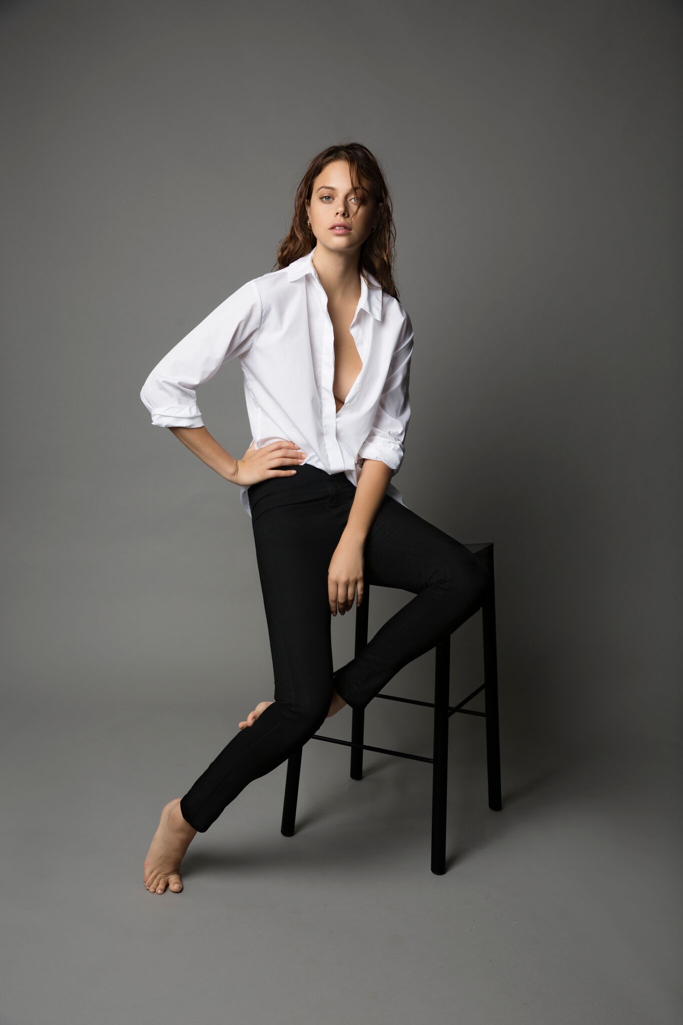 Brunette model in white shirt sitting on black stool against grey background
