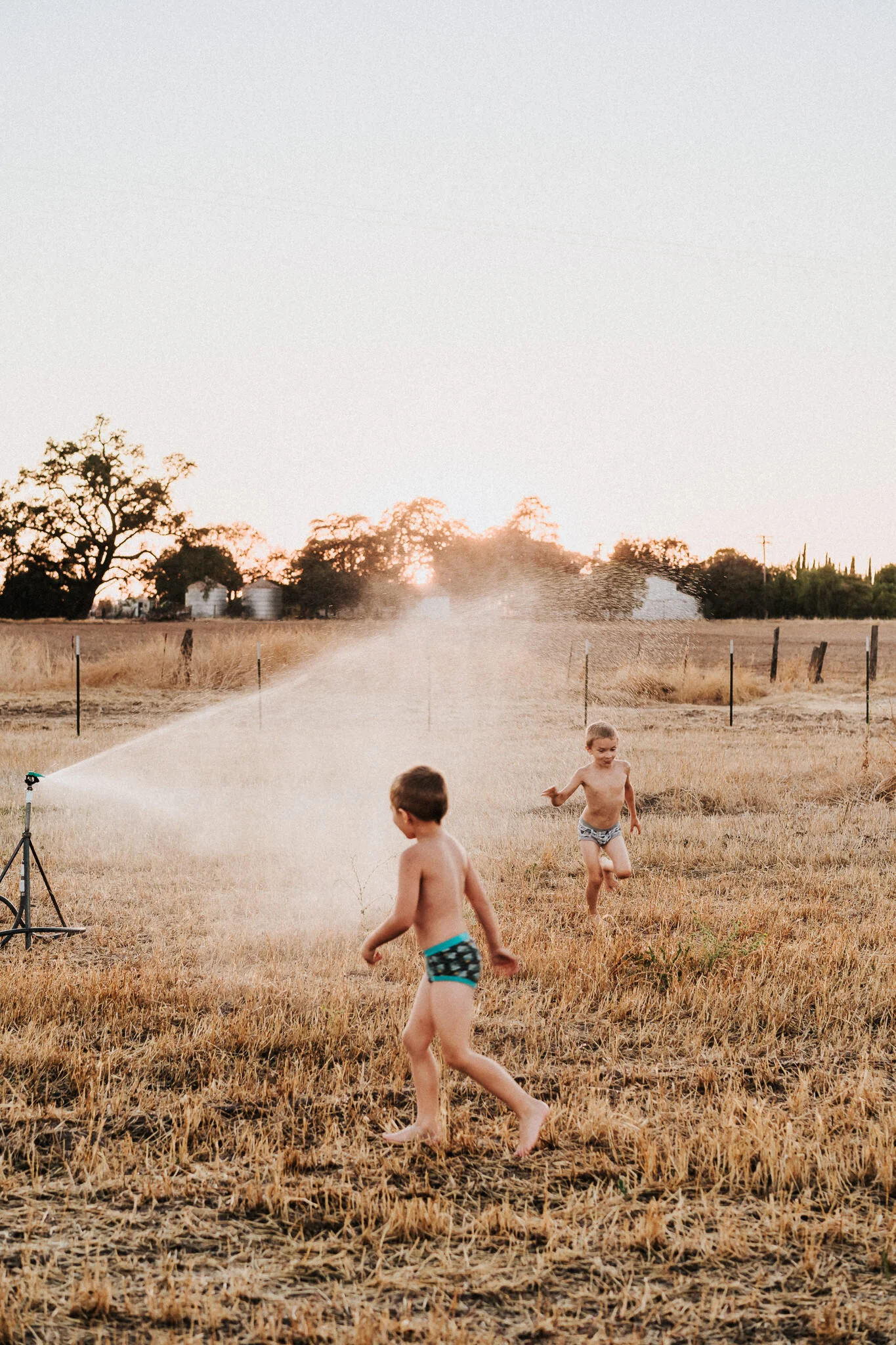 Two boys run around the sprinkler.