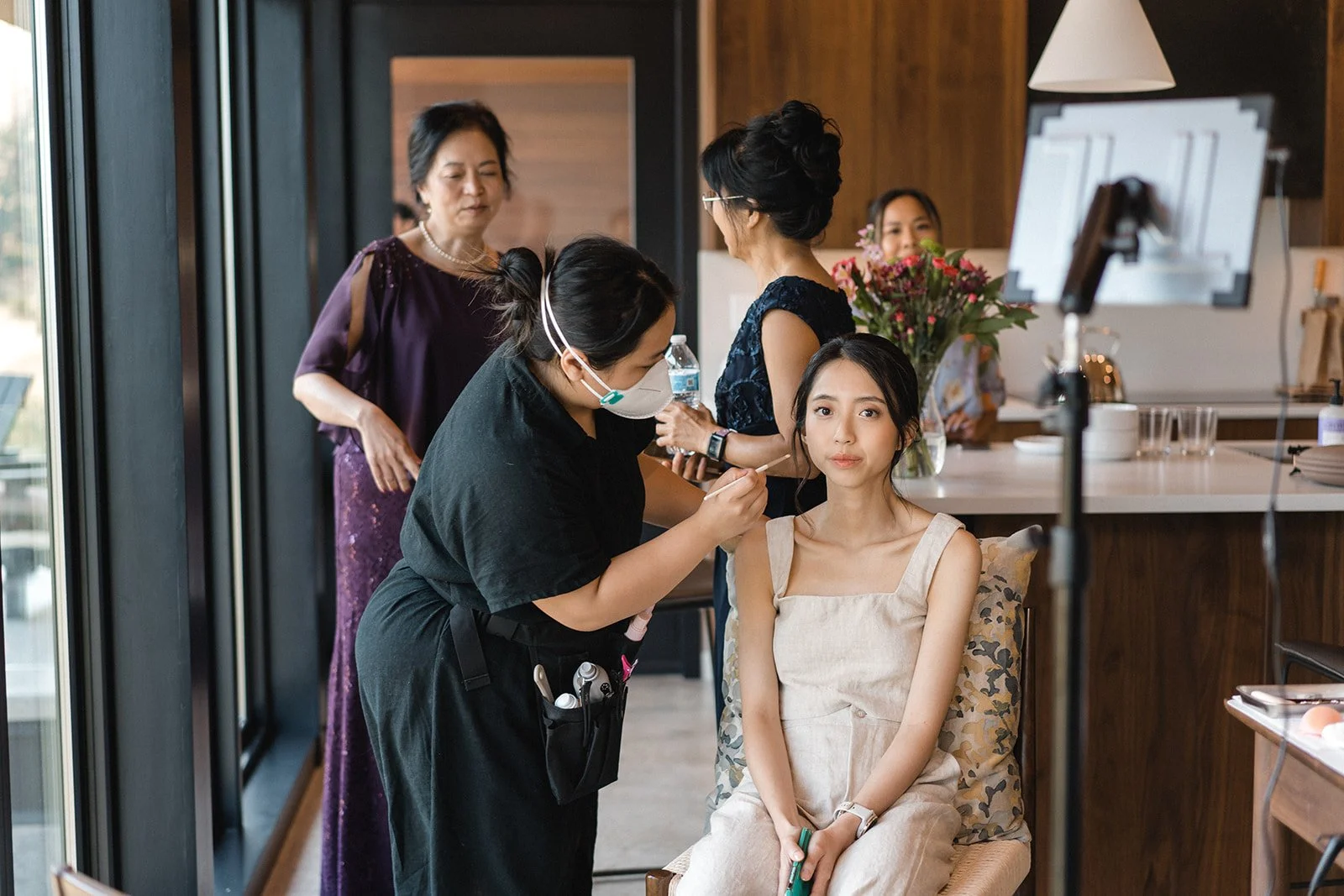 Bride looks towards the camera as make-up artist brushes the bride's face with blush.