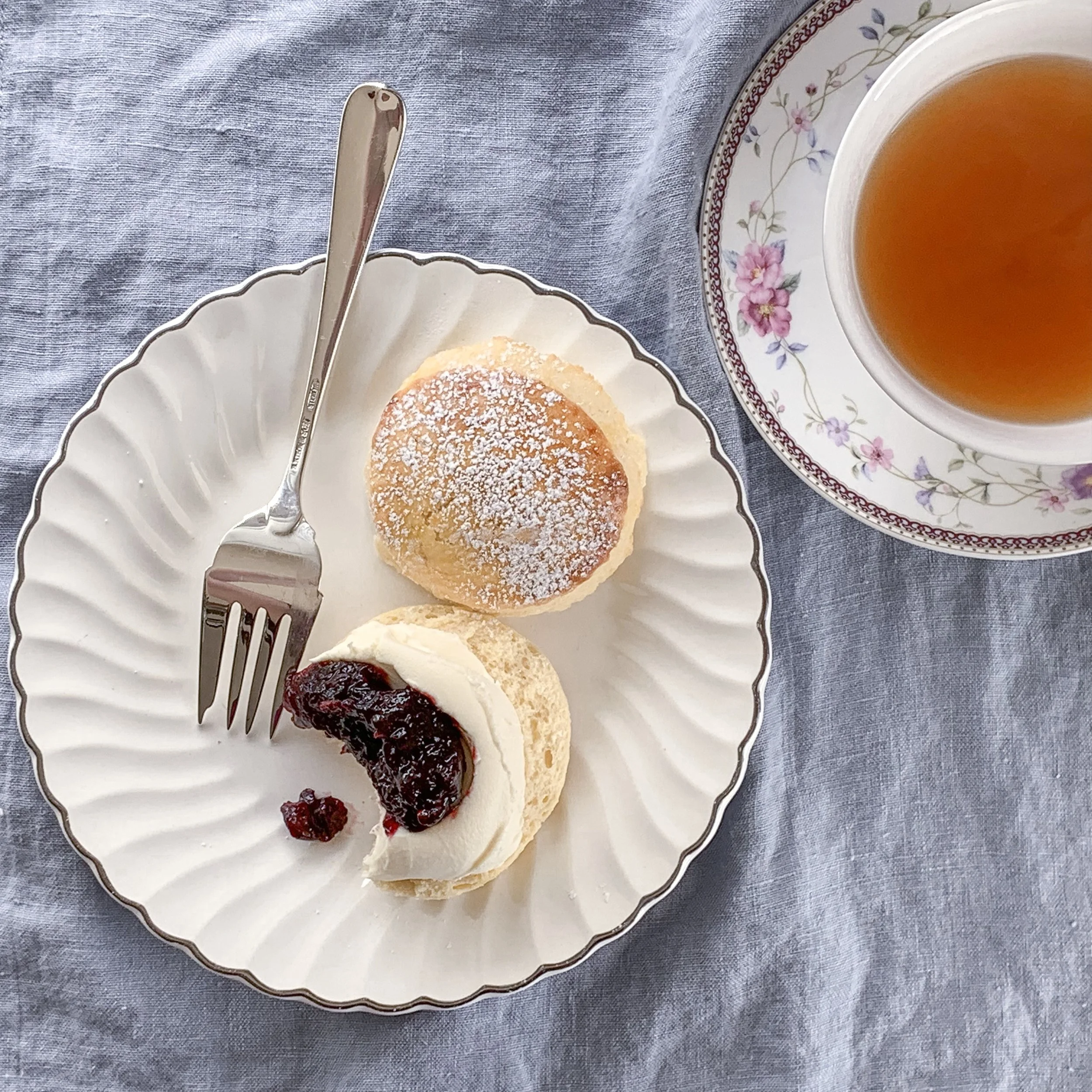 Tea in a teacup with a plate that has a Plain British style scone with cream and jam in the Devonshire style. Cream first, jam second.  On a blue linen table cloth