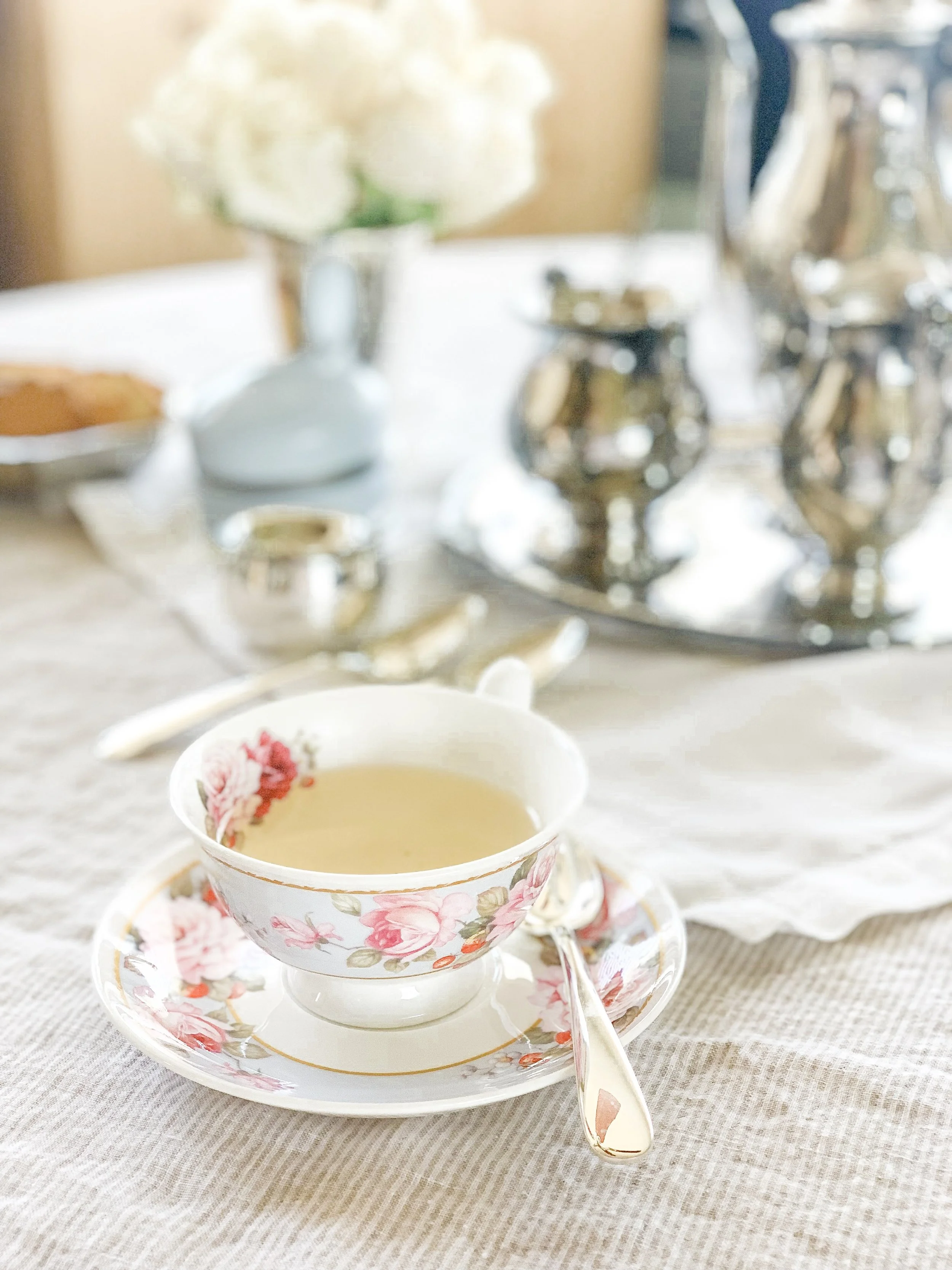 A teacup in the foreground with white tea in it. A silver tea set is out of focus in the background