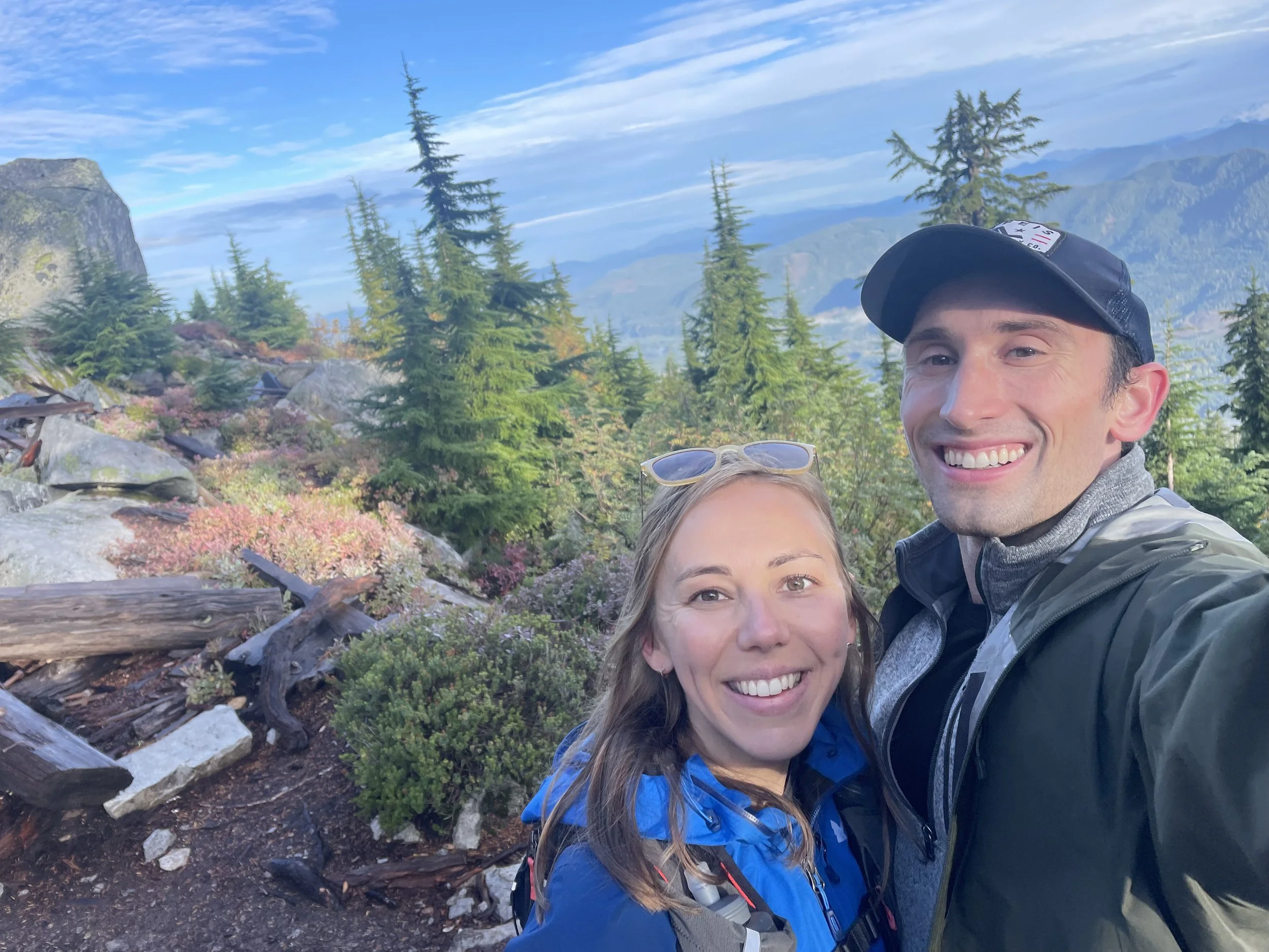 blonde woman has sunglasses on her head. Man has baseball cap. They are on a trail overlooking seattle and are smiling in the selfie.