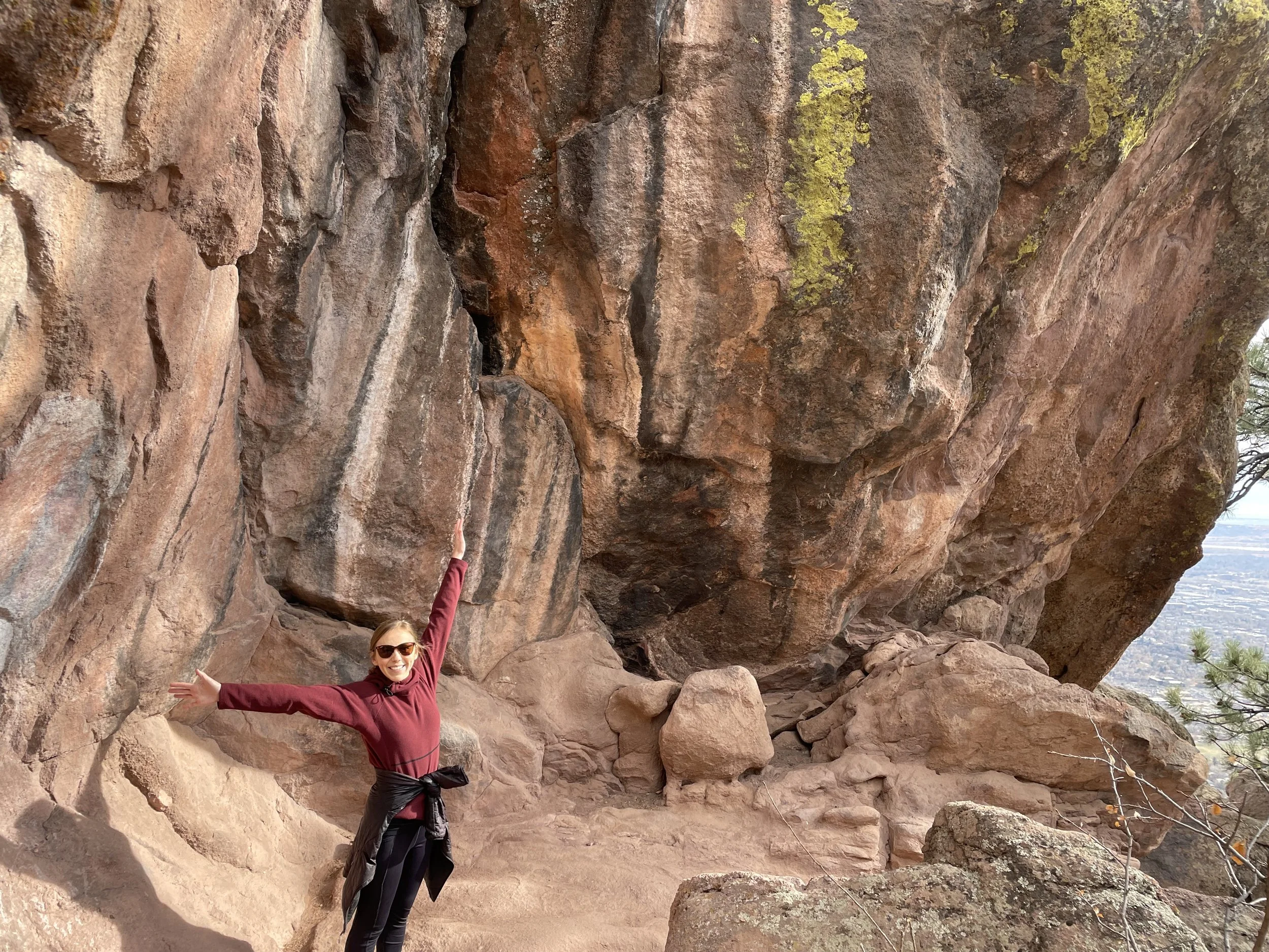 Dr. Brynn Olinger is standing on a trail in the PNW. She is wearing a burgandy sweater, her arms are wide open and she is smiling