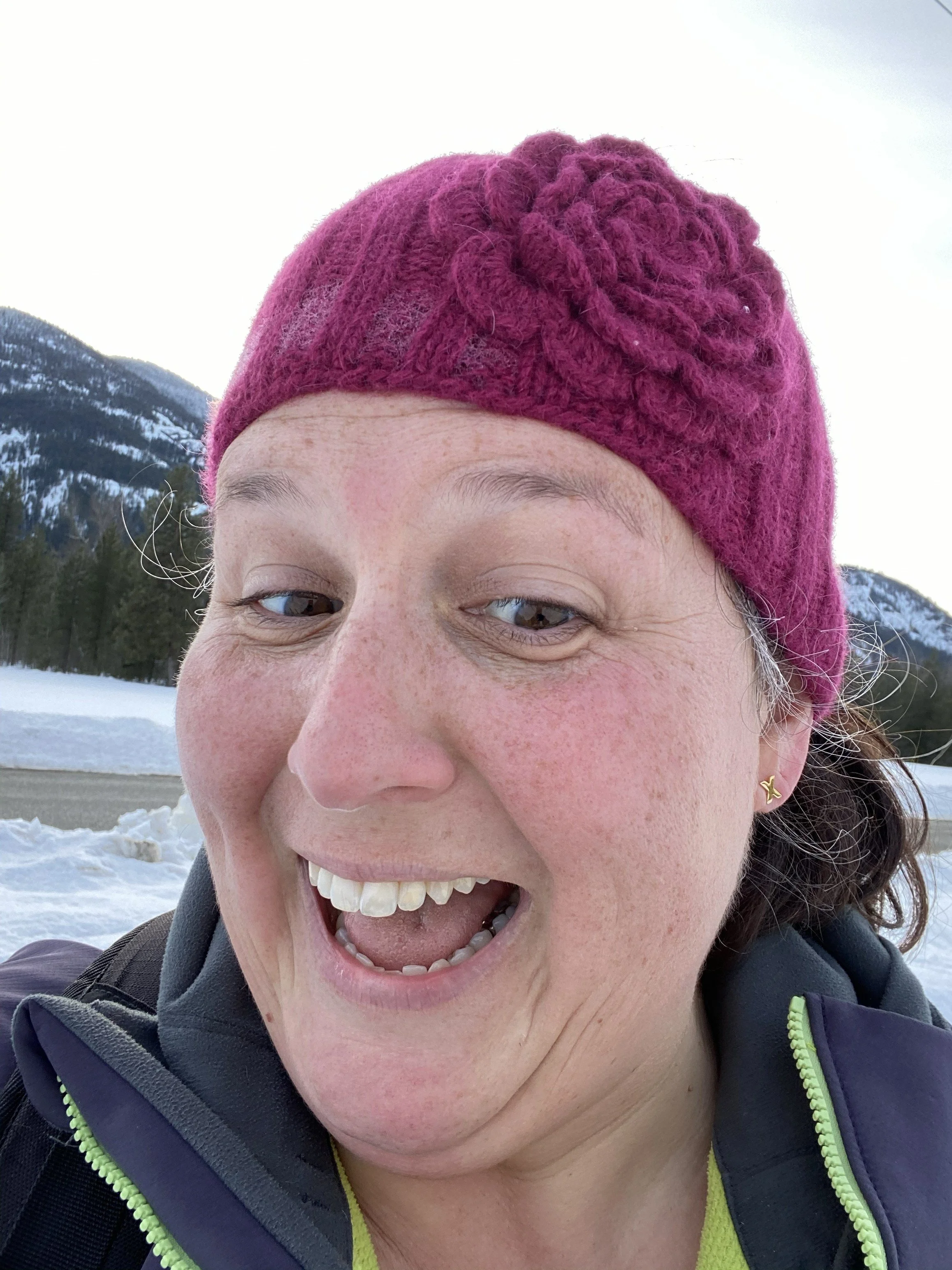 woman wearing maroon beanie with flower crochetetd on it is skiing and smiling with wide smile