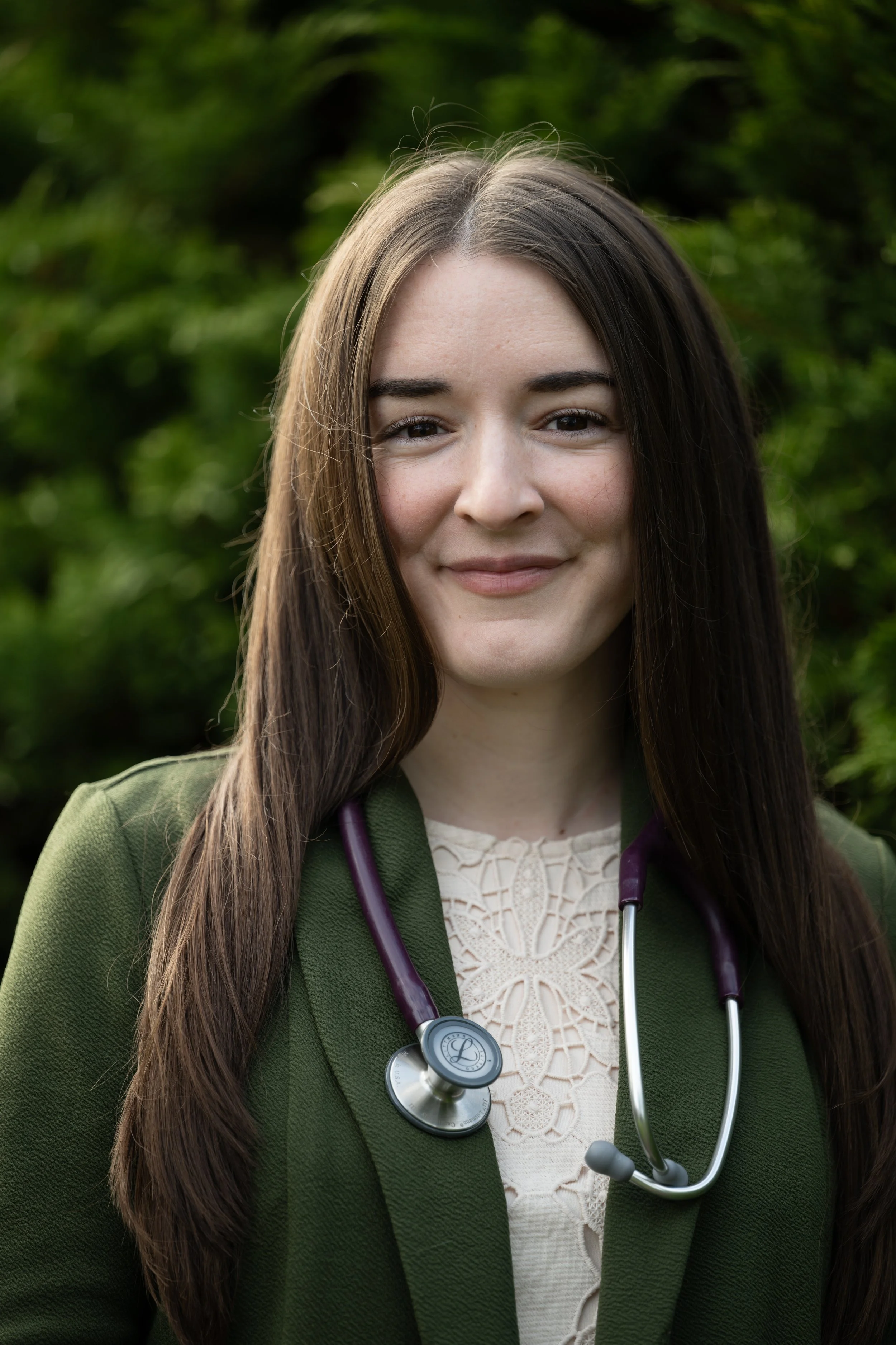 Dr. Claire Hannes is standing in front of greenery. Her brown hair is long. She is wearing a green blazer and a stethoscope