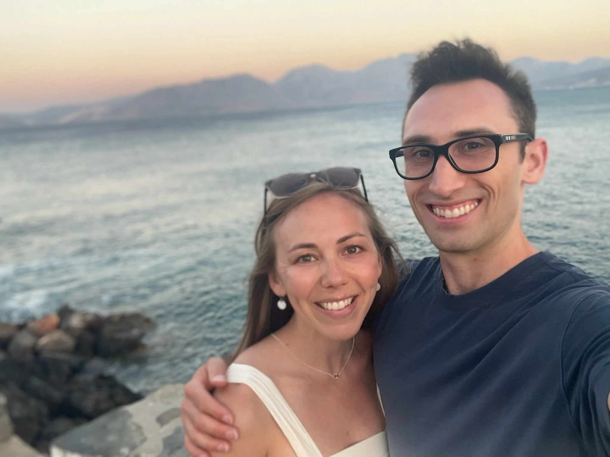 blonde woman and dark haired man stand arm in arm with seattle's coast behind them. There is a foggy sunset behind them.