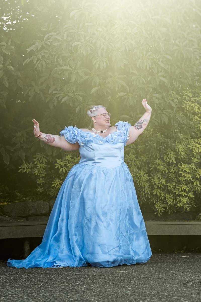 woman dressed in blue ball gown smiles and has arms out up towards the sky, joyful
