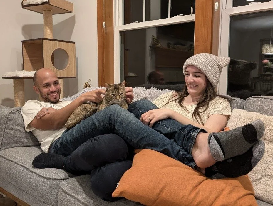 man and woman sit on a couch and are smiling at the cat on their lap. A cat tree is in the background.