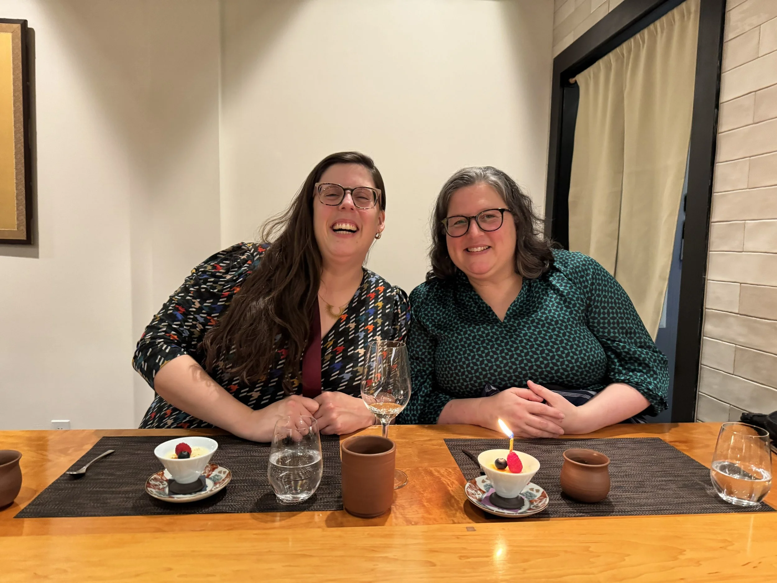 two women sit at sushi restaurant table. a candle is lit in a dessert in front of one of the women. Both are smiling.