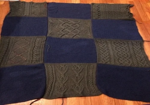 Image shows afghan as of August 18, 2015. It is composed of three strips of 4 squares each. 8 of those squares are blue seed stitch, 8 of those squares are green (the light is very yellow saturated, but I promise they're green) in various cable patterns.