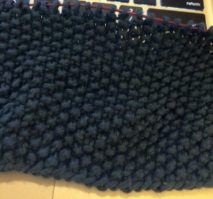Image shows one of the plain squares in progress. It is a dark blue yarn, knit in seed stitch.