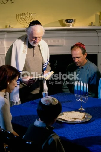 Image shows an older balding man wearing a tallit and holding an open siddur standing next to a table. To his left is a seated woman with short hair, to her right is a young boy in a sweater vest, button down shirt and Hanukkah themed suede kippah. To the right of the older man is a seated man in a long sleeved polo shirt with a red satin kippah. There is someone seated next to this man, but the image cuts off everything but some arm in a blue sleeve. On the table is a silver plate with square machine made matzah and an unlit ḥanukkiyah with all of the arms holding blue and white striped candles.