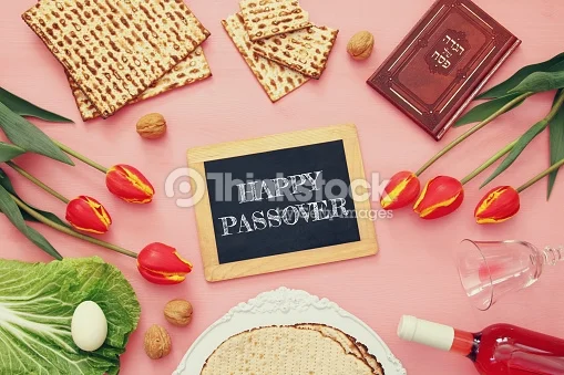 Image shows a wood framed slate with "HAPPY PASSOVER" written on it in stylized chalk lettering. Surrounding the frame clockwise from noon are: pieces of square machine made maztah, un-shelled walnuts, a red-brown haggadah , red tulips with yellow stripes, a wine glass on its side with a splash of wine still left, a bottle of pink wine on its side, a stack of three round hand made matzahs on a white cloth, more un-shelled walnuts, a leaf of lettuce with an egg on it, and more tulips.