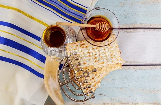 Image shows a tallit with blue and gold stripes over white painted wooden slats. Positioned over the tallit are (clockwise from top): glass bowl with honey and wooden dipper, two broken sheets of square machine made matzah, a ḥanukkiyah lying flat on its side, a shofar , and a silver kiddush cup partially filled with wine and sitting on a silver coaster.