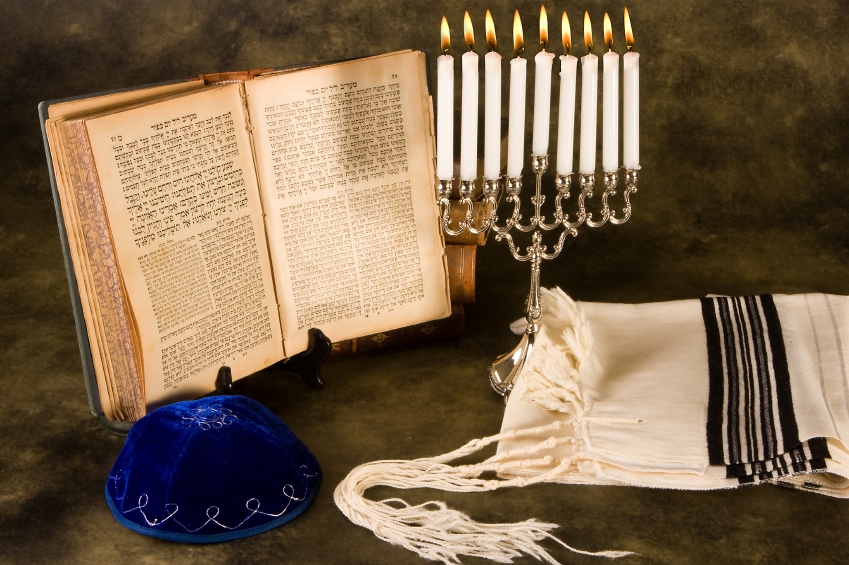 Image shows an open Hebrew prayerbook on a stand, a blue velvet kippah with silver embroidery, a ḥanukkiyah with all its candles lit, and a folded tallit with black and silver stripes.