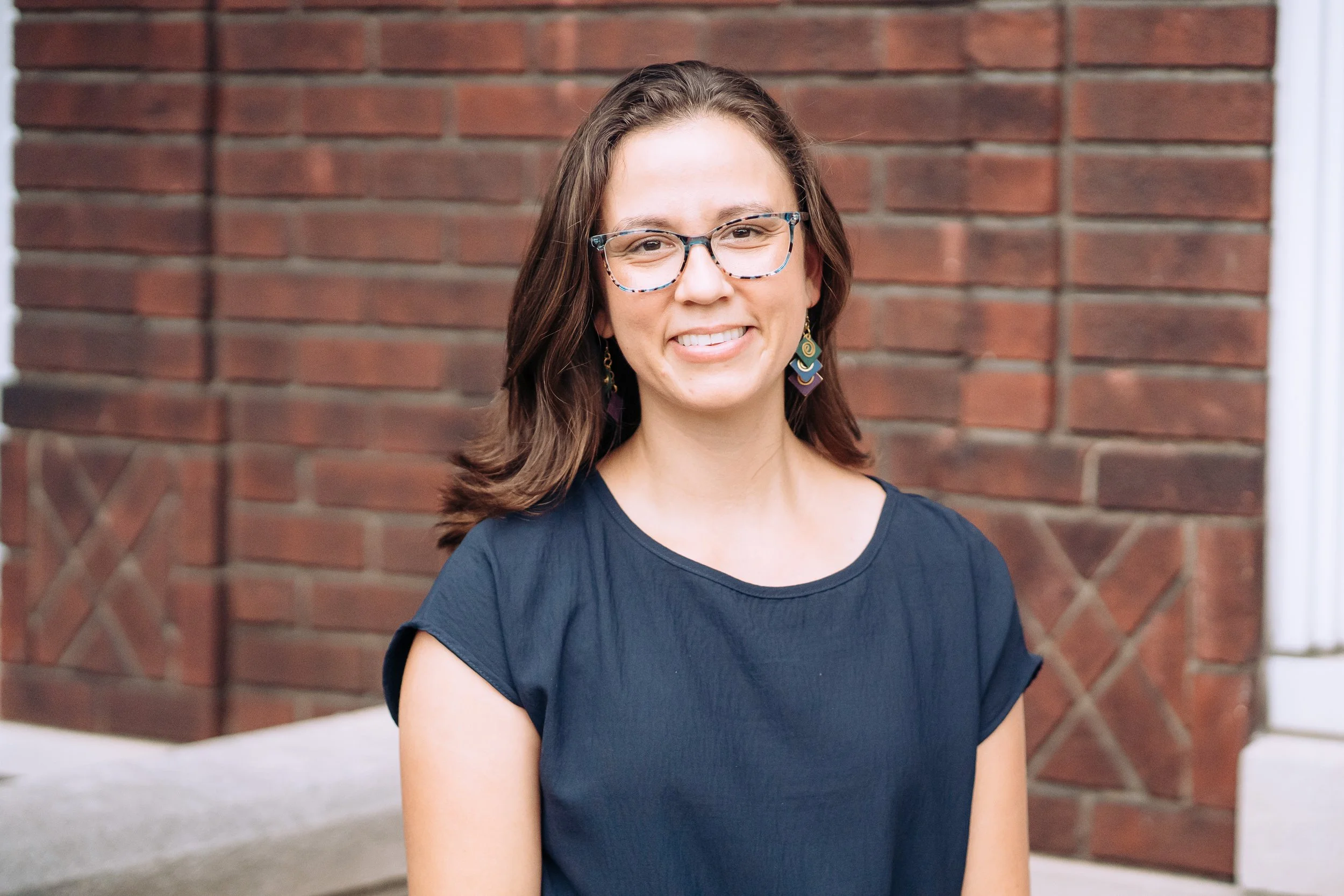 A woman with shoulder-length brown hair, wearing glasses and earrings, smiling while standing outside in front of a brick wall.