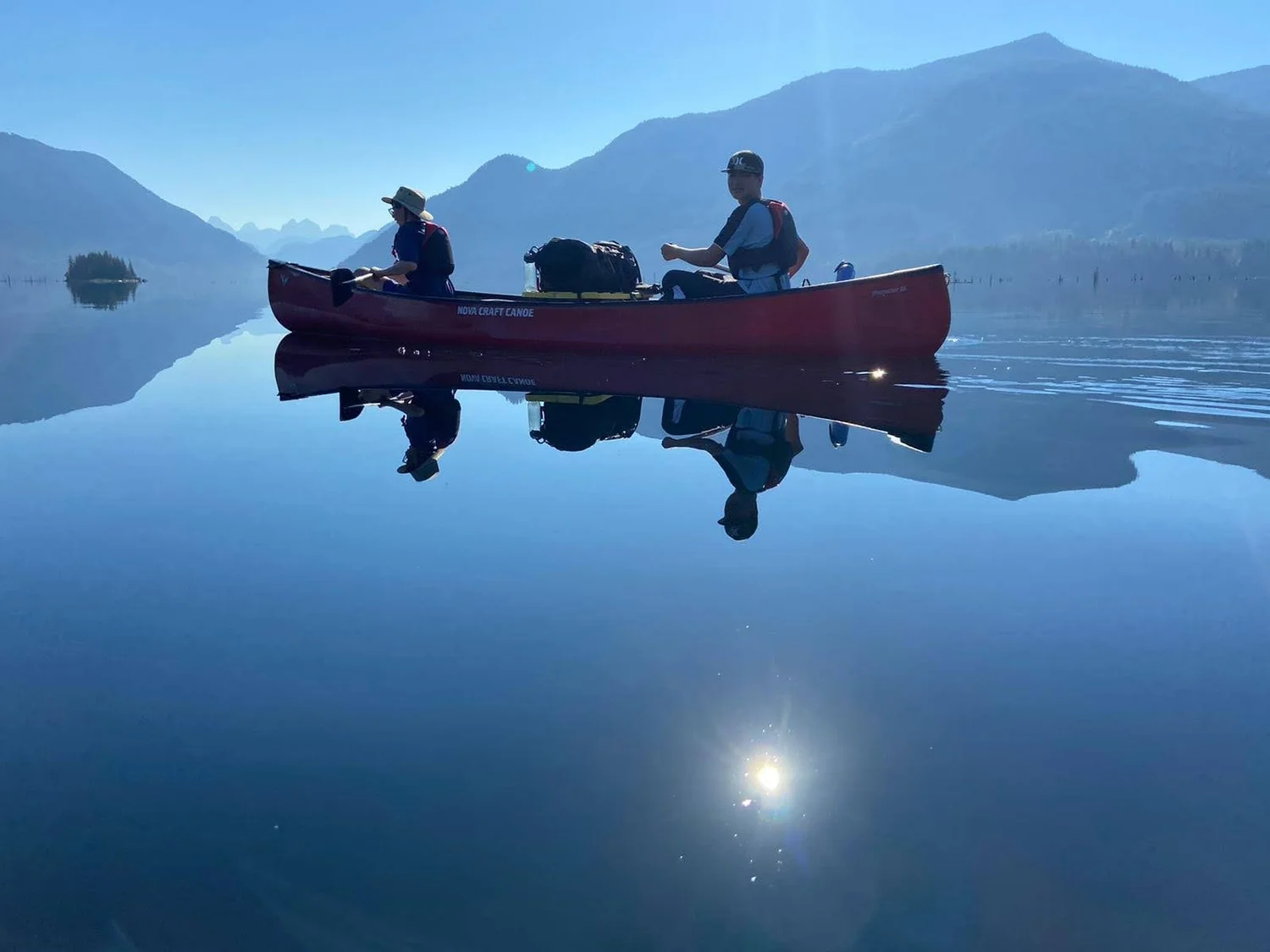 Two paddlers in a loaded red canoe reflected in still water beneath the mountains