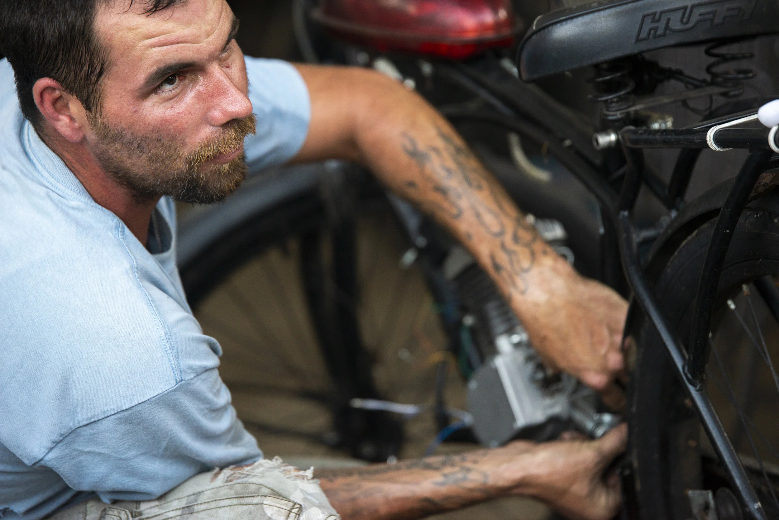 Calvin Kirby looks onward at an interaction between two people conversing in his backyard while working on a motorized bicycle.
