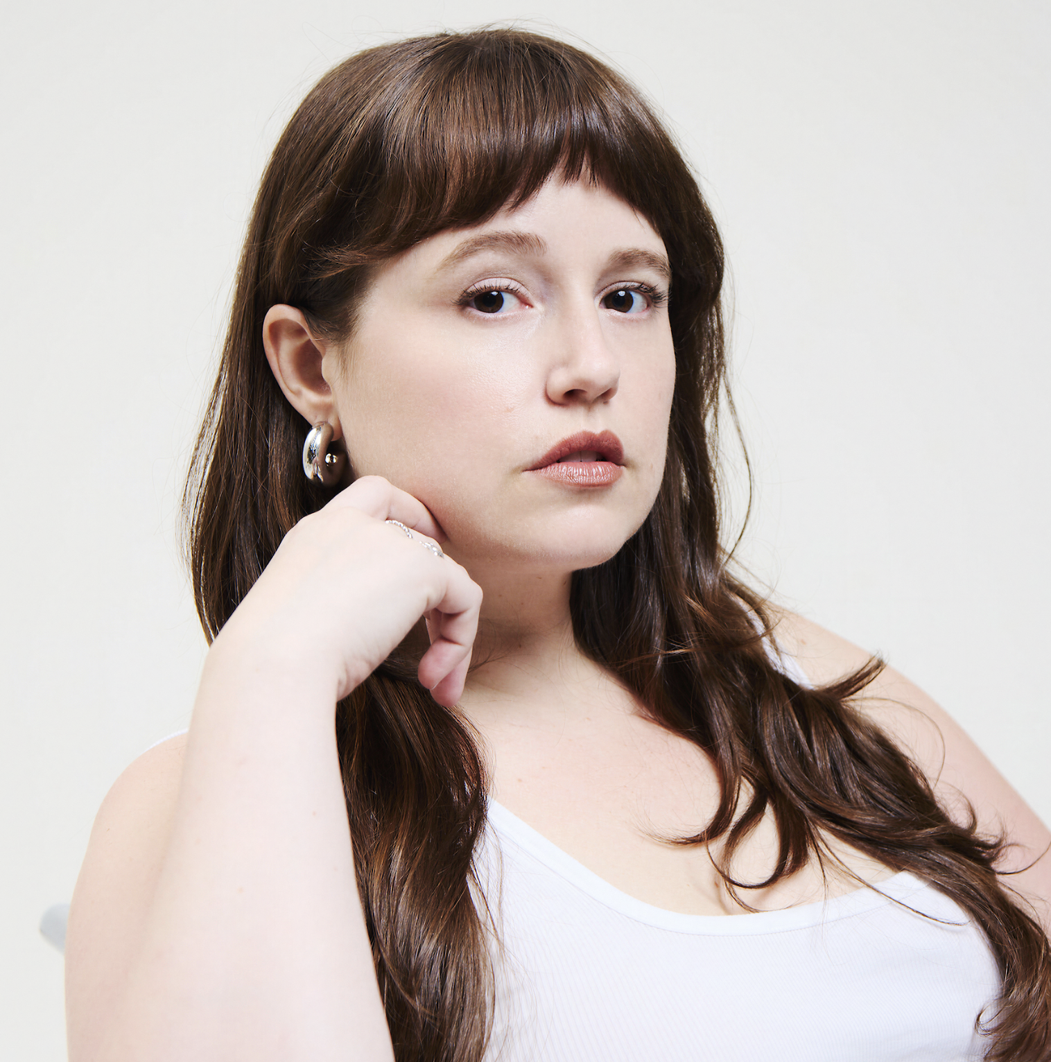 Close-up portrait of a woman with long brown hair and bangs wearing silver hoop earrings and a white tank top.
