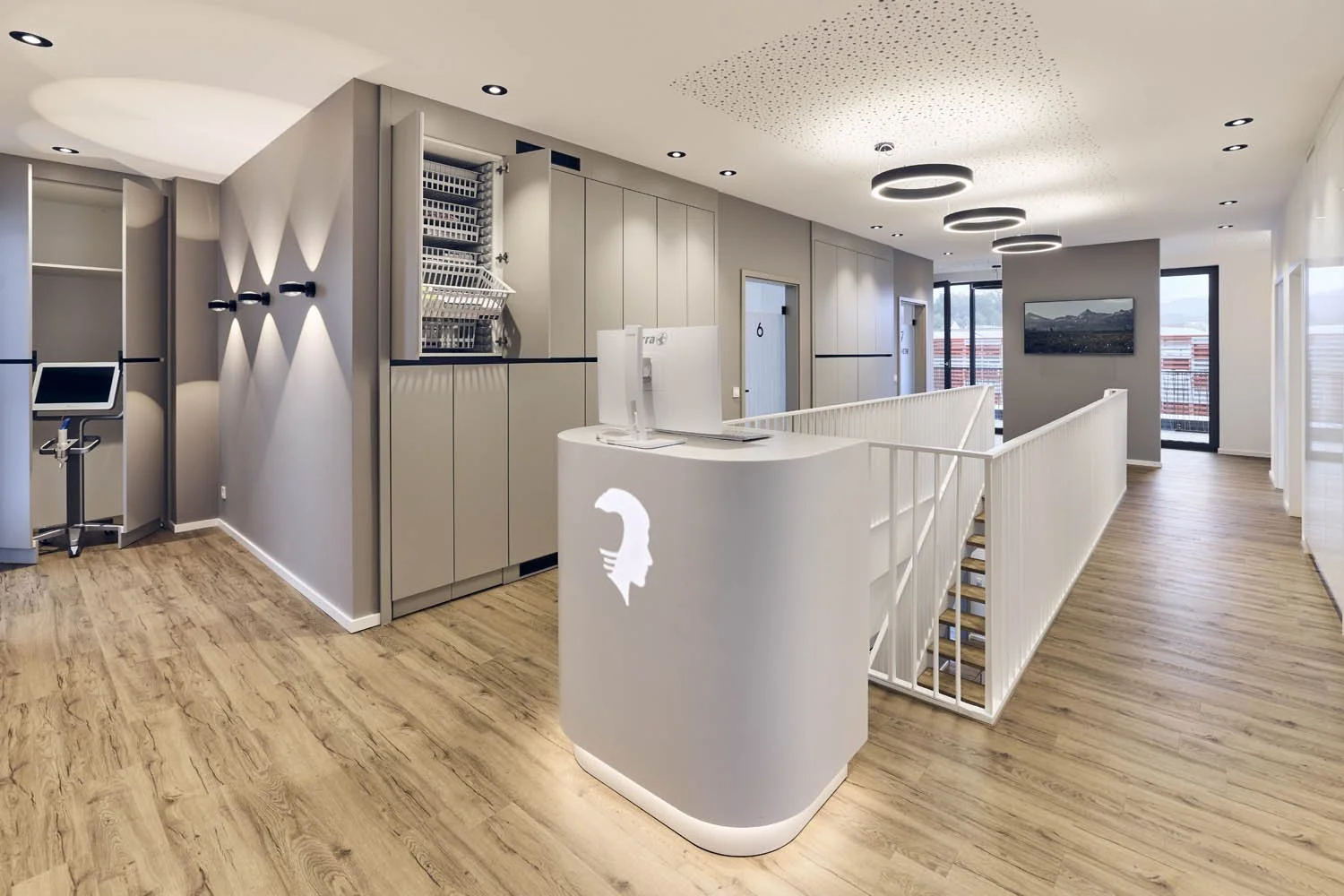 Moderne, helle Rezeption in einem Bürogebäude mit modernem Design, grauen Wänden, Holzfußboden, Computer auf dem Schreibtisch, und Blick auf Fenster mit Balkon im Hintergrund.