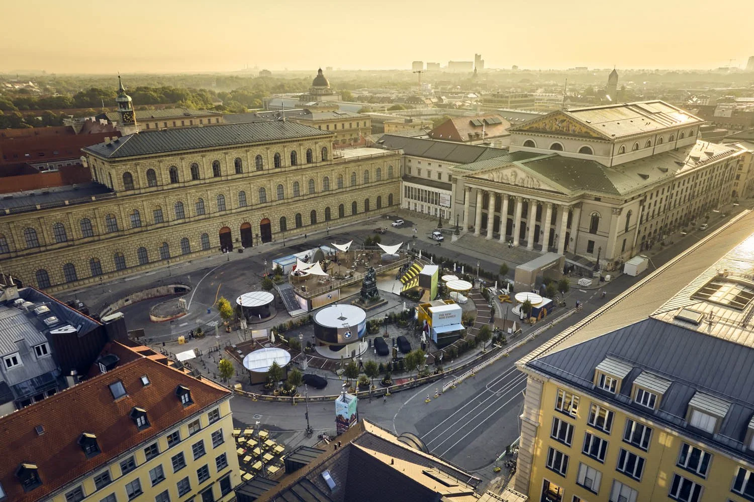 Luftaufnahme eines historischen Stadtplatzes mit Gebäuden im neoklassizistischen Stil, einem Platz mit Zelten und Pavillons, und mehreren Touristen, bei Sonnenuntergang in München, Deutschland.