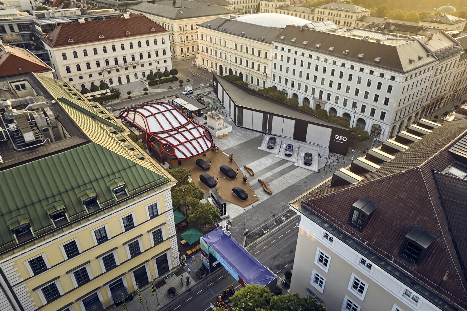 Luftaufnahme eines Stadtplatzes mit mehreren historischen und modernen Gebäuden, einem roten und weißen futuristischen Dach, Parkplätzen und einer Audi-Ausstellungshalle.