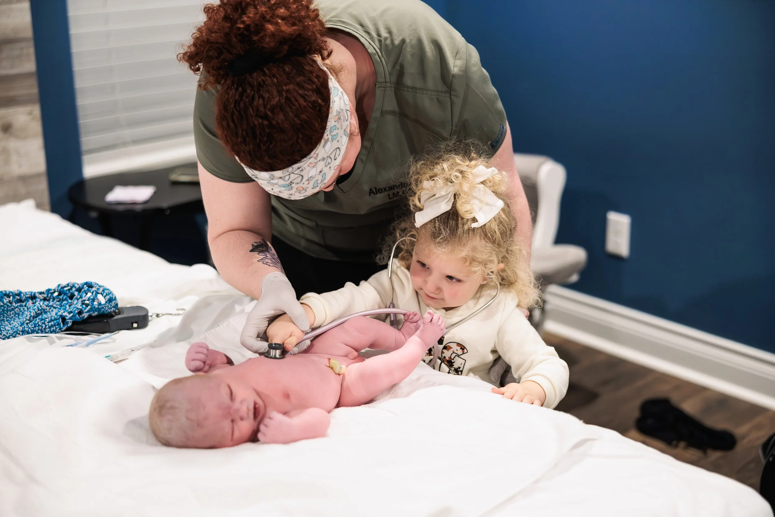 Midwife Alex performing a newborn baby check in a birth center, while letting the new big sister listen to her brothers heart beat with a stethoscope.
