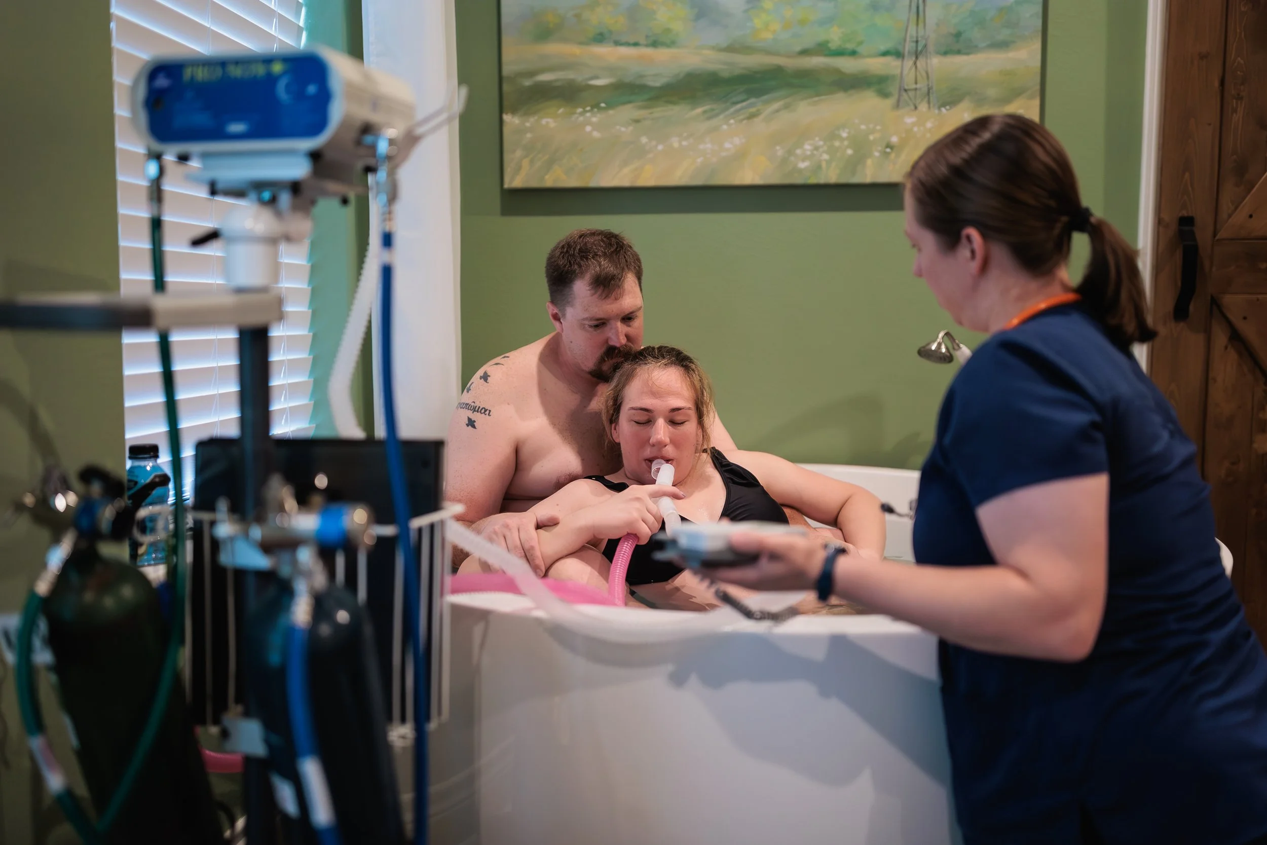 A mom laboring in a birth tub with her husband supporting her, while using nitrous oxide for pain relief, and a nurse birth assistant listening to baby heart tones with a doppler.