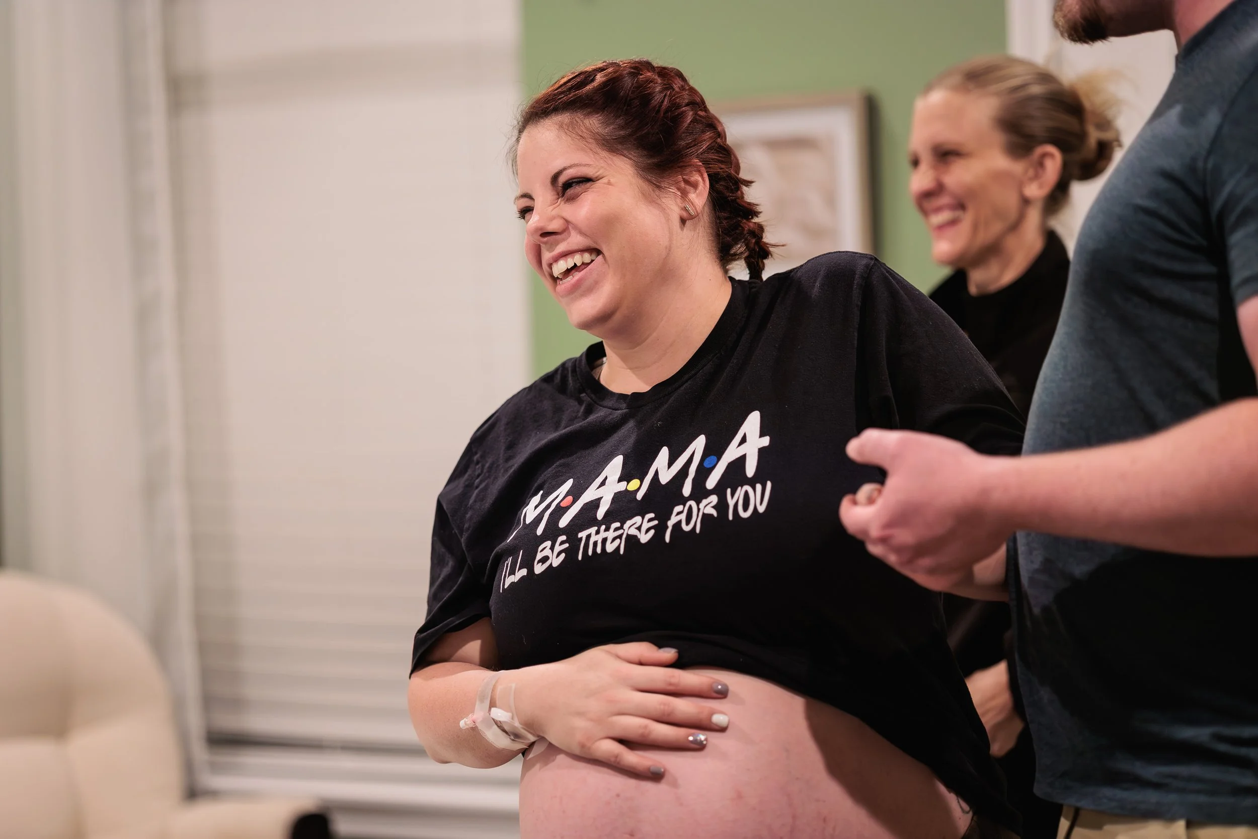 A woman in early labor wearing a black T-shirt with the text 'MAMA I'll Be There For You' is smiling and laughing while holding her pregnant belly, at the Heritage Birth Center.