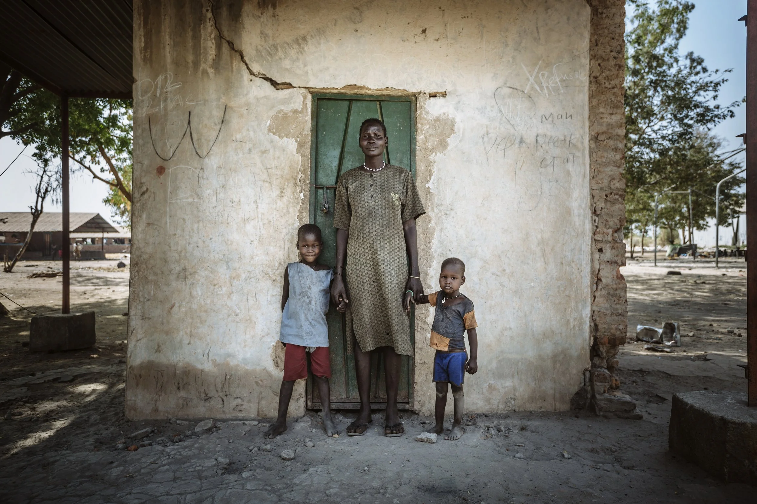 A woman stands with her two children outside a square building in Bentiu Town, South Sudan. Formerly attached to a larger building, the square room, which is all that remains now, is where this young family has been living for the past six months.