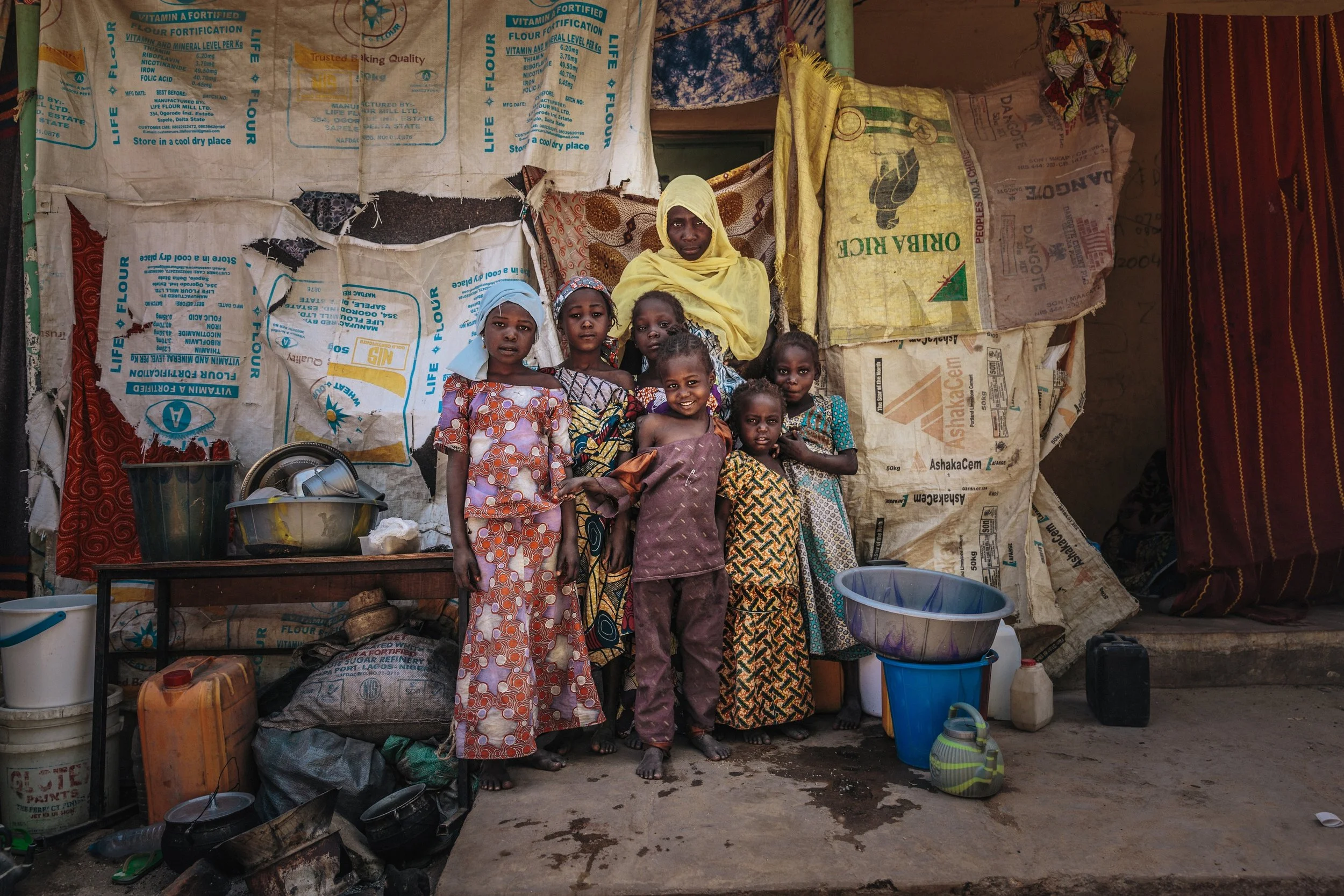 Atai and her children pose in front of their makeshift shelter in the one of the IDP camps in Maiduguri, Nigeria. It is largely constructed from burlap sacks. Atai had lived with her family, working as a tailor, in her village, Bama. Just over two years ago, Atai and her children fled when Boko Haram attached the village. Boko Haram kidnapped her daughter and killed her husband during the brutal attack.