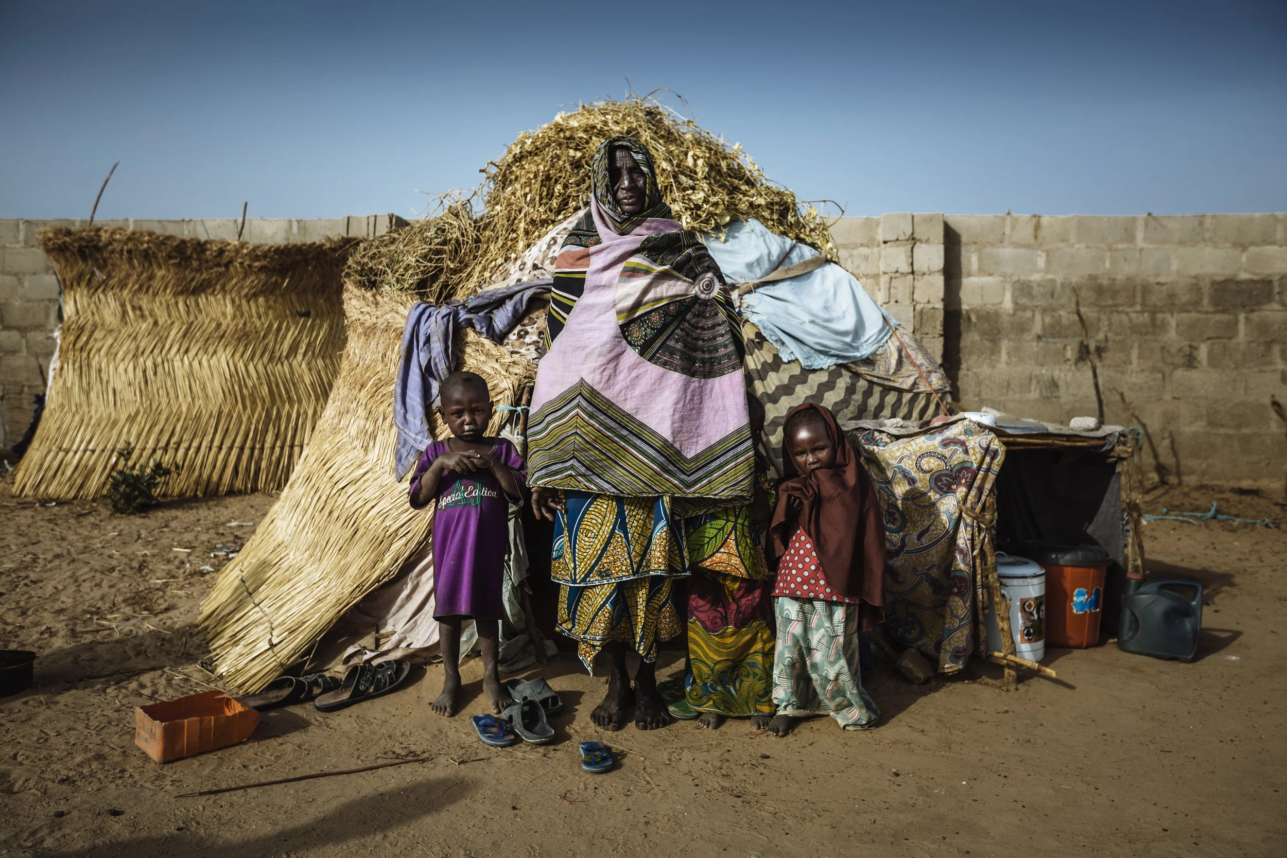 A woman stands with her grandchildren outside of the small straw house, where they all live. The family fled their village months earlier, when it was attacked by Boko Haram, and now live in a host community in Maiduguri, Nigeria.
