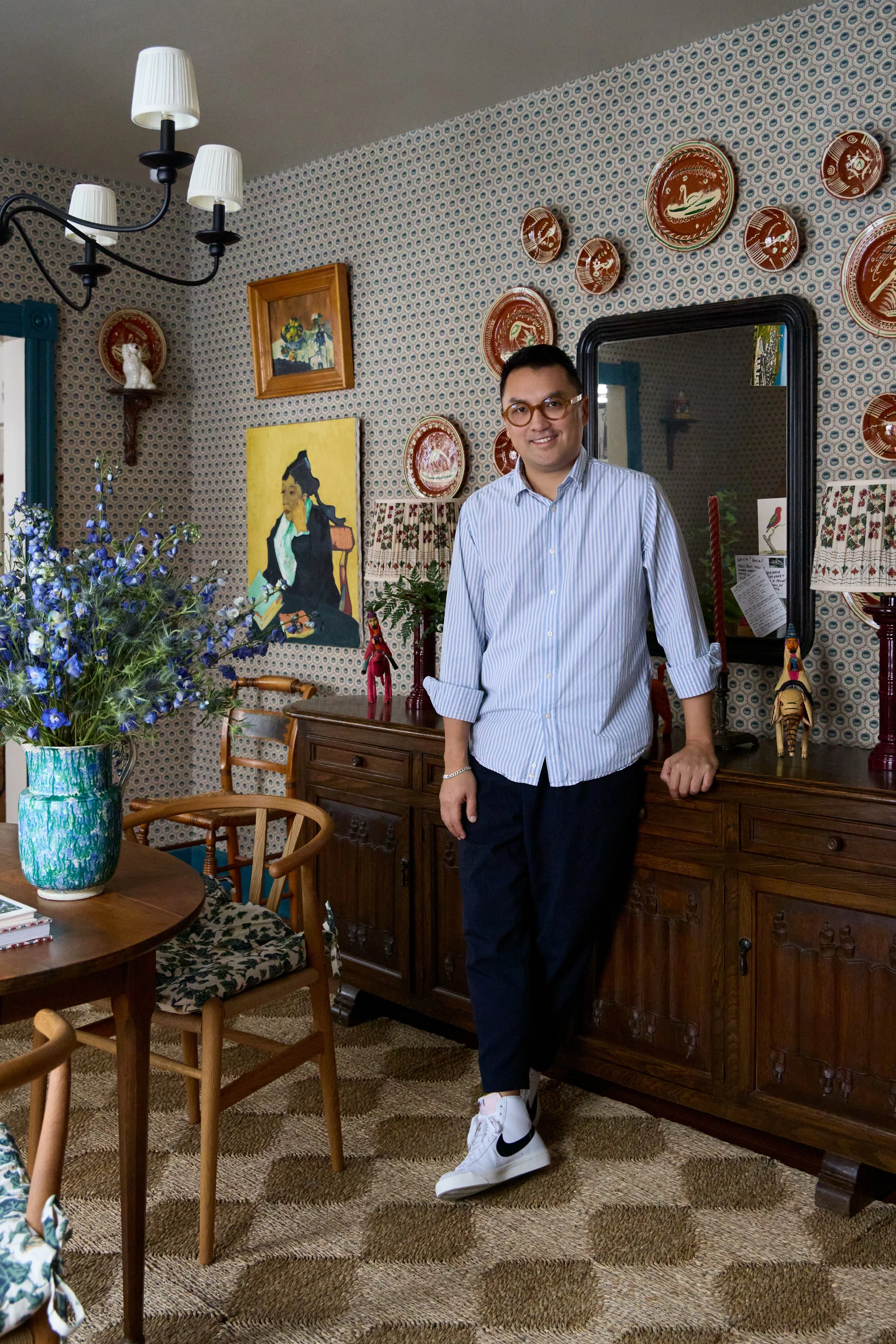 A man wearing glasses in a striped shirt and dark pants, standing in a decorated room with vintage furniture, artwork, and floral wallpaper.