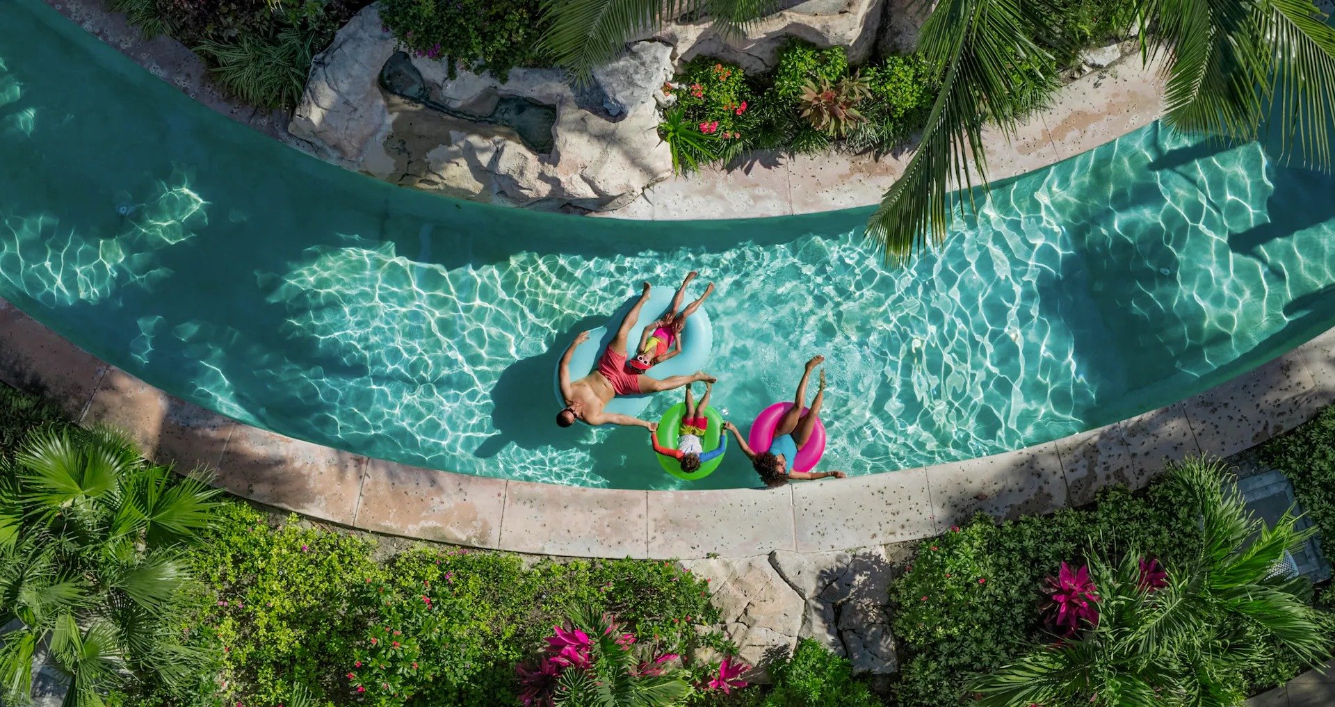 Three boys riding waves on boogie boards in a water park, with water splashing around them and a rocky structure in the background under a blue sky.