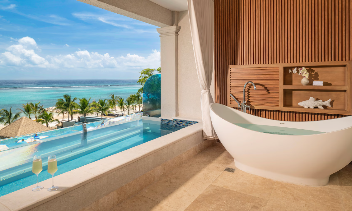 Clear turquoise water at a tropical beach with white sand, palm trees, beach chairs, thatched umbrellas, colorful buildings, and a red-roofed structure on an island in the distance under a blue sky with scattered clouds.