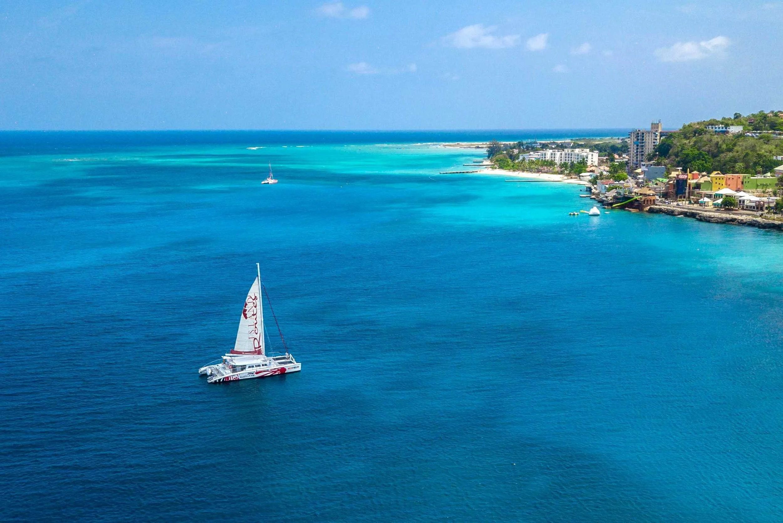 A catamaran sailboat with a large sail labeled 'RE/MAX' floating on clear turquoise water near a lush green island.