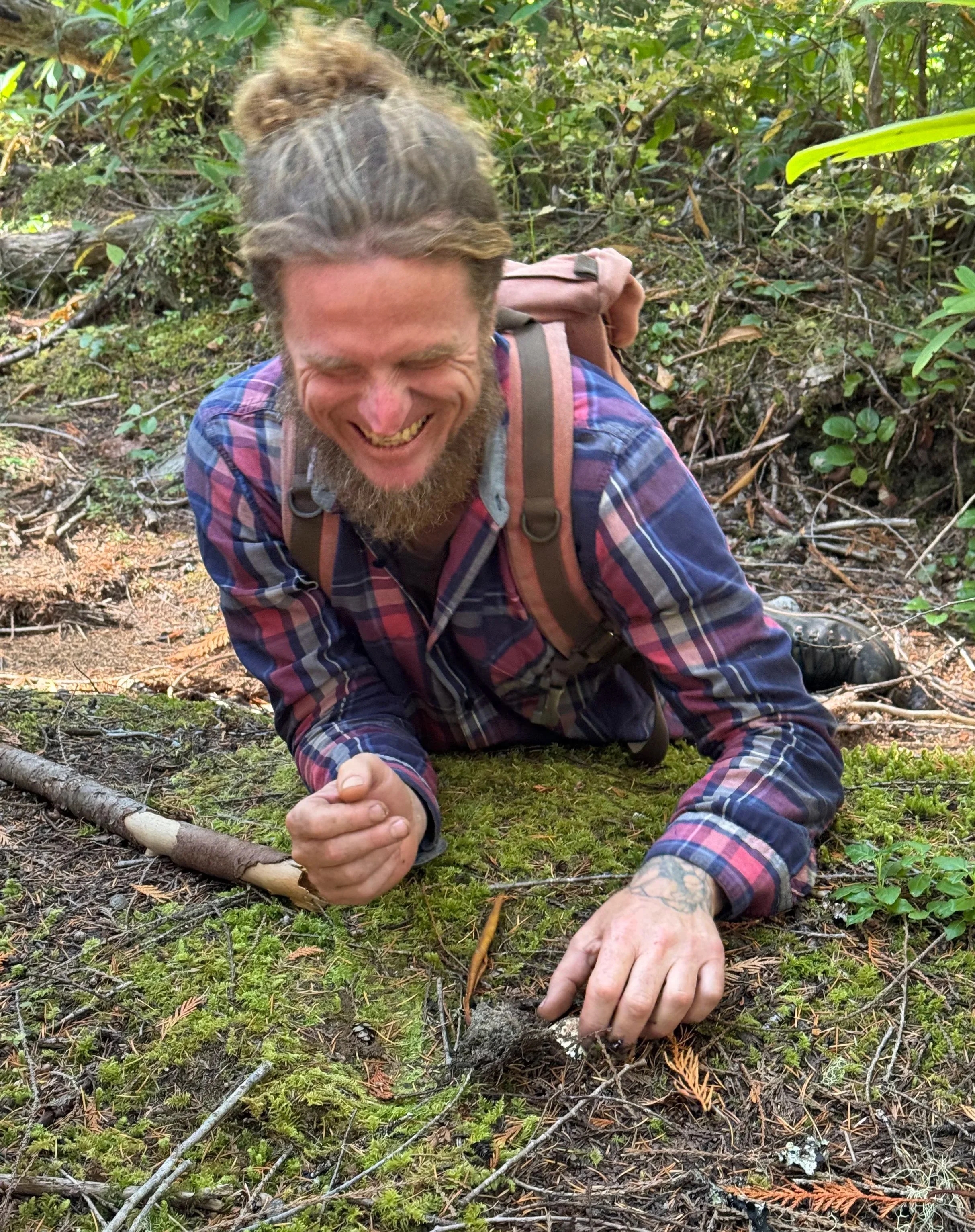 a person in a plaid shirt and backpack laughing while trying to unearth a matsutake mushroom from the ground