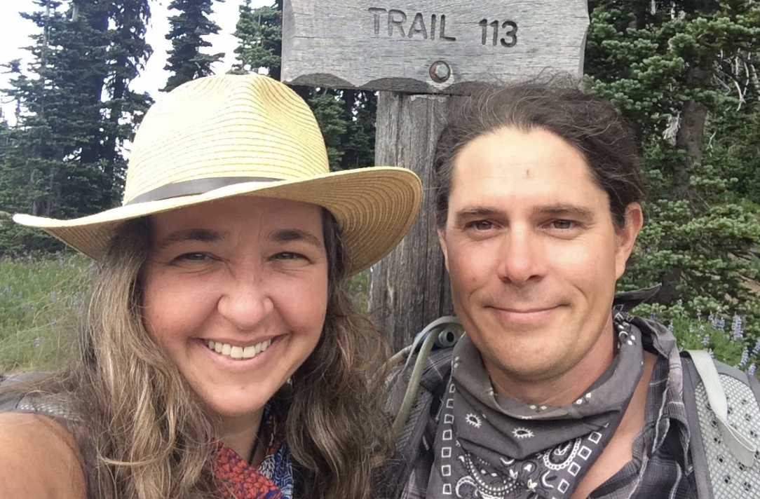 A woman and man taking a selfie outdoors near a trail sign that reads 'TRAIL 113', with trees and greenery in the background. The woman is smiling and wearing a wide-brimmed hat, while the man is smiling and wearing a bandana around his neck.