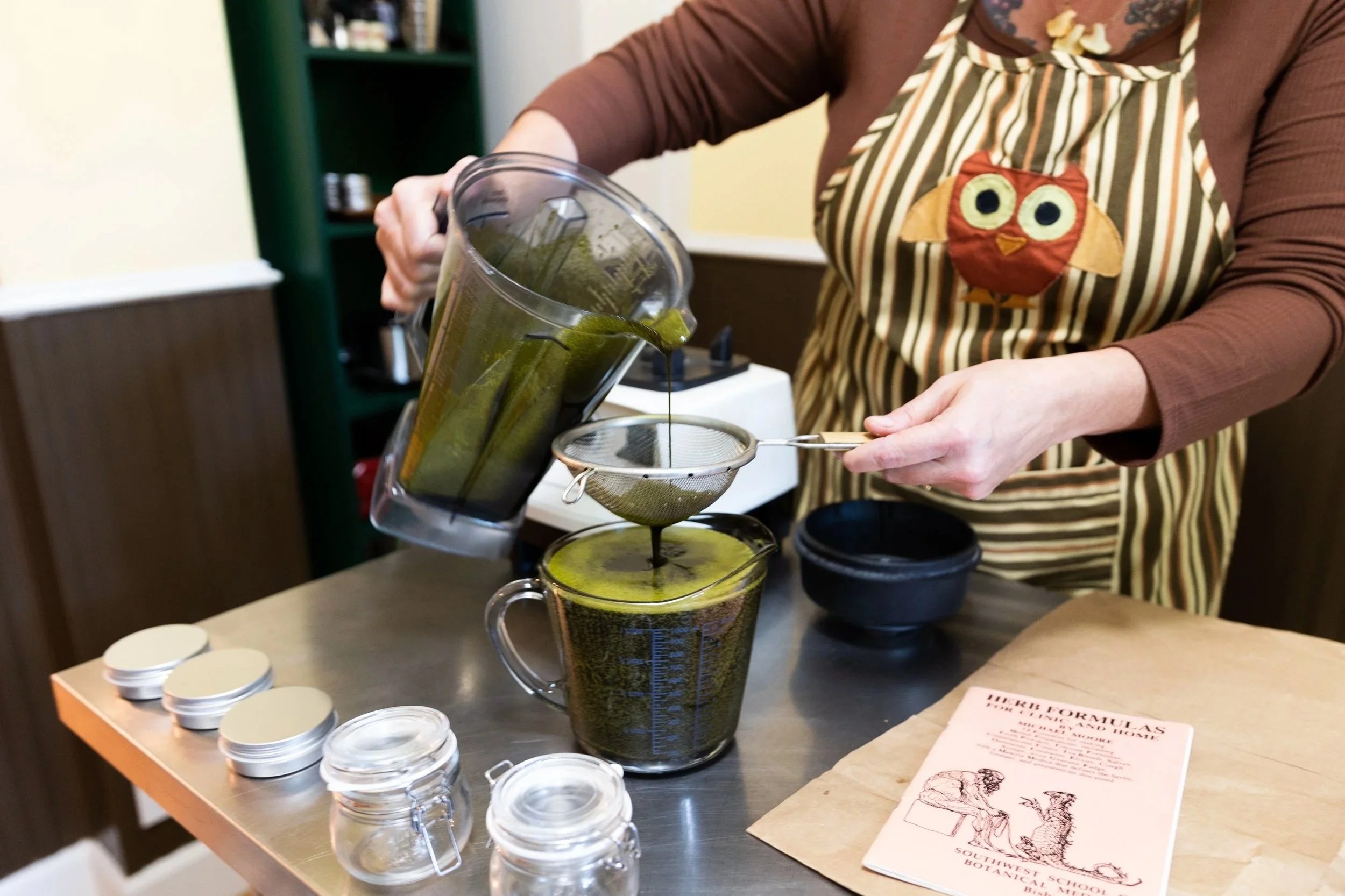 A person pouring bright green liquid from a blender into a glass measuring cup, with several small jars with metal clips on a table. The person is wearing a striped apron with an owl design and a brown long-sleeved shirt.